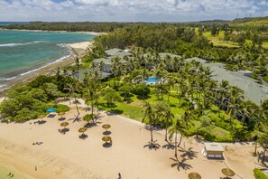 An aerial view of a resort with a beach, palm trees, and a swimming pool.