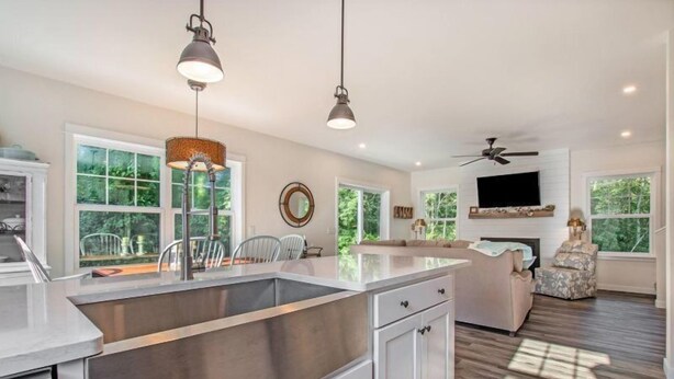 Una cocina amplia con una isla grande, armarios blancos e iluminación colgante. Hay un área de comedor con un sofá y sillas, y un televisor montado en la pared.