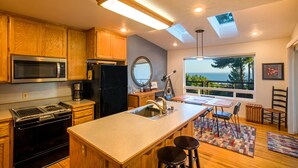 A modern kitchen with wooden cabinets, a built-in microwave, a refrigerator, and a large island with a sink. There is a dining area with a table and chairs, and a large window offering a view of the ocean. - Wow! Amazing Oceanview, walk to Beach & Game room! (Whitethorn)