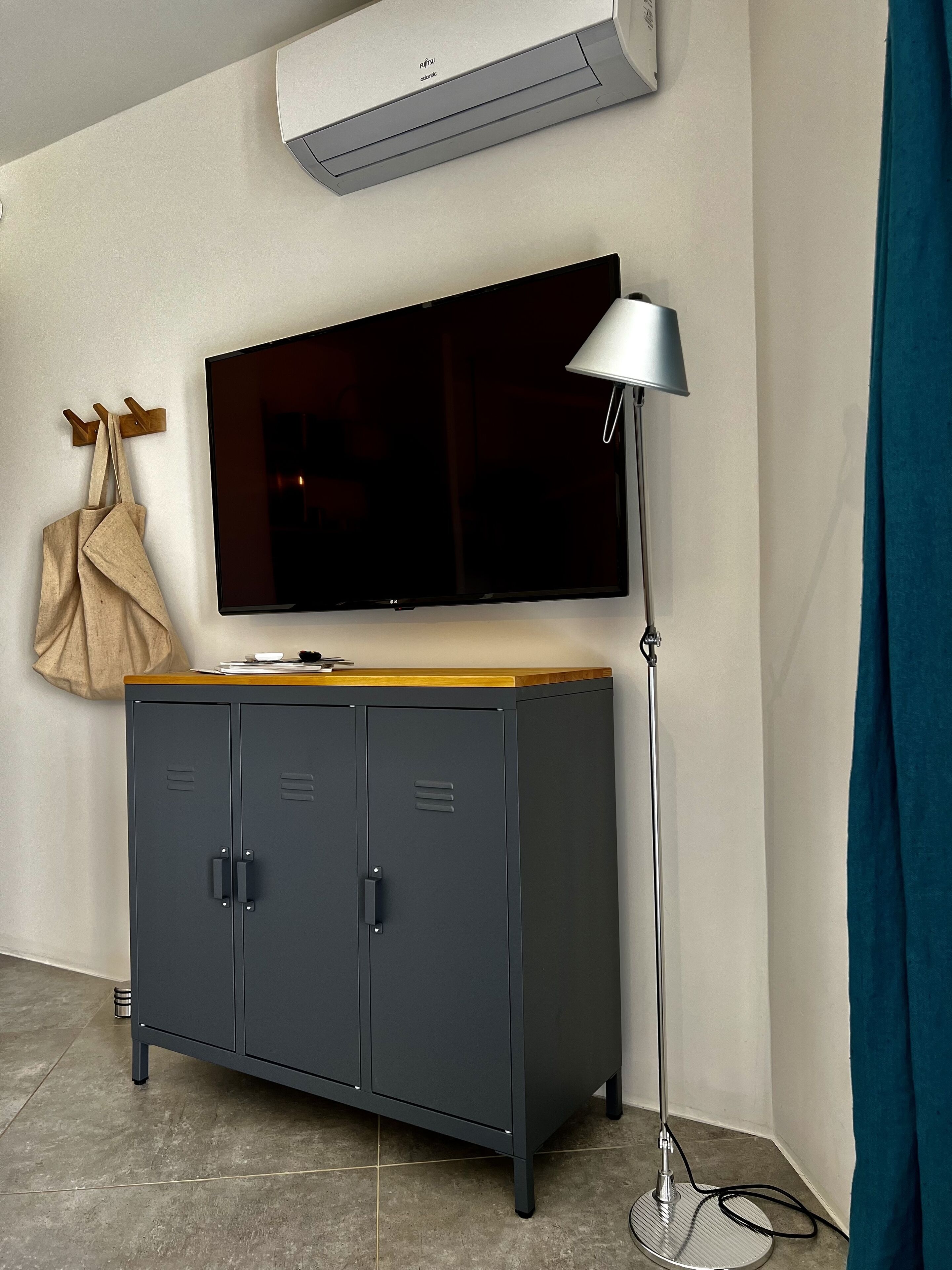 Et værelse med et mørkegråt skab, et fladskærms-tv ophængt på væggen, en gulvlampe og en beige taske hængende på en træbøjle.