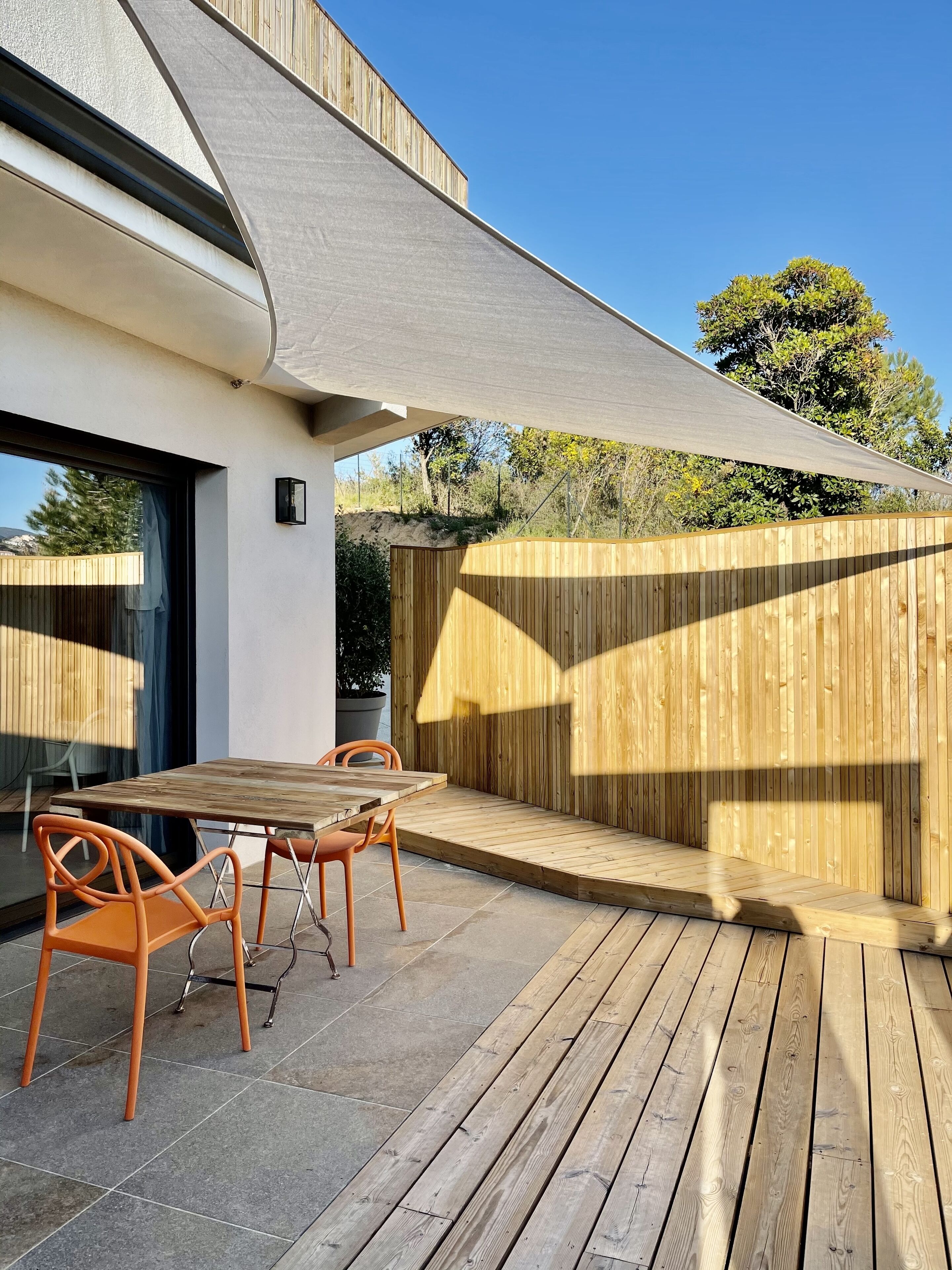 An outdoor patio with a wooden table and chairs, a wooden deck, and a large shade sail.