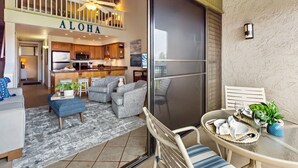 Un vaste espace de vie comprenant un coin cuisine, un balcon avec une table et des chaises, et une vue sur la cuisine, le tout agrémenté d’une inscription « ALOHA ».
