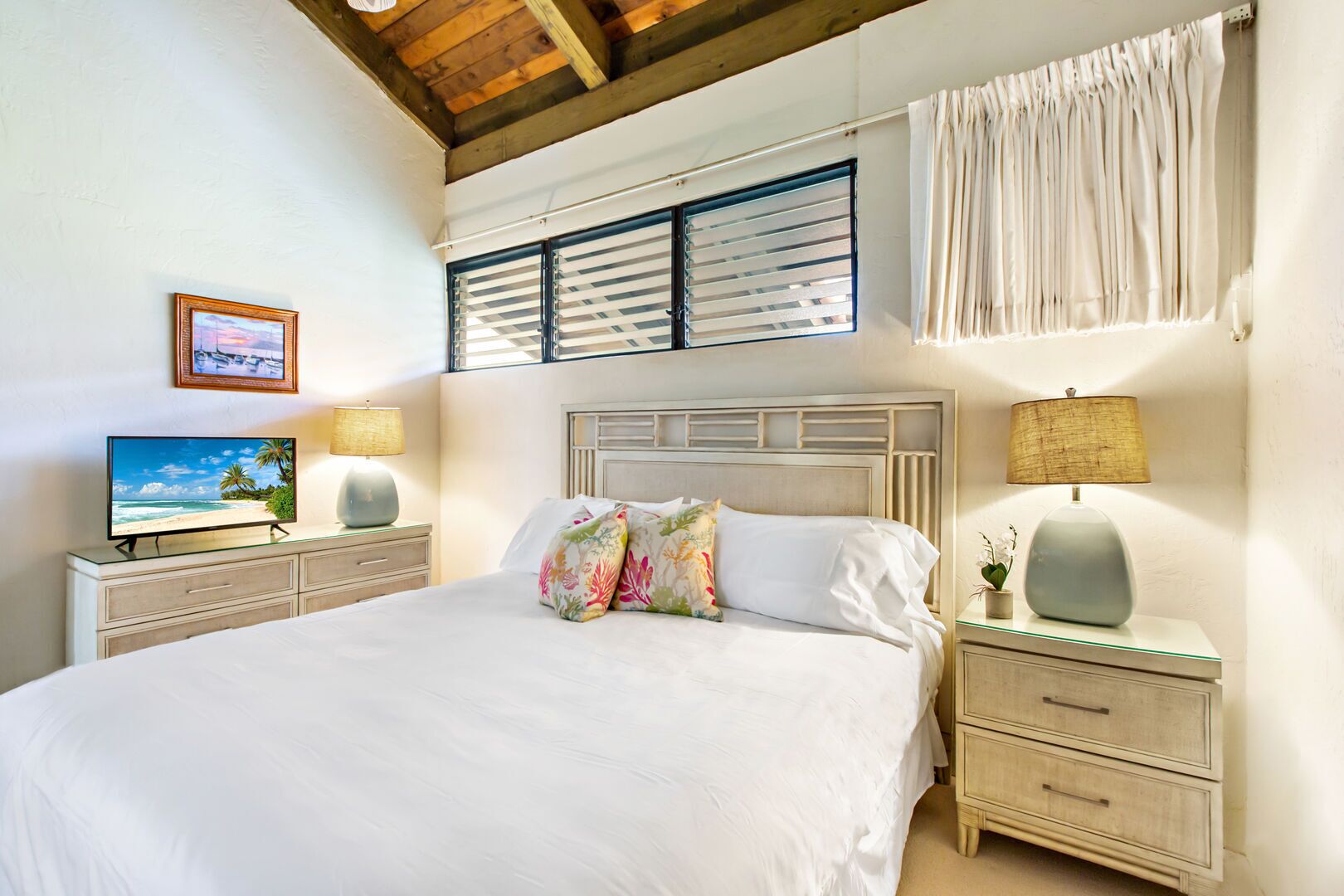 Une chambre avec un grand lit, deux tables de chevet avec des lampes, un téléviseur posé sur un meuble, un tableau encadré au mur et une fenêtre avec des volets.