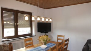 A dining area with a wooden table, chairs, a vase with flowers, a blue placemat, and a flat-screen TV mounted on the wall.
