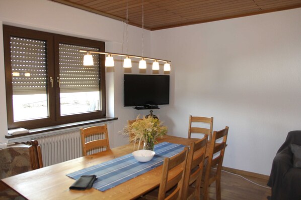 Un comedor con mesa de madera, sillas, un jarrón con flores, un camino de mesa azul y un televisor de pantalla plana montado en la pared.