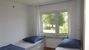A room with two single beds, each with a blue pillow and a white mattress. There is a window with white curtains, and a radiator is visible below the window.