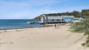 A beach with a pier extending into the water, a large blue house with a balcony, and a sandy shore.