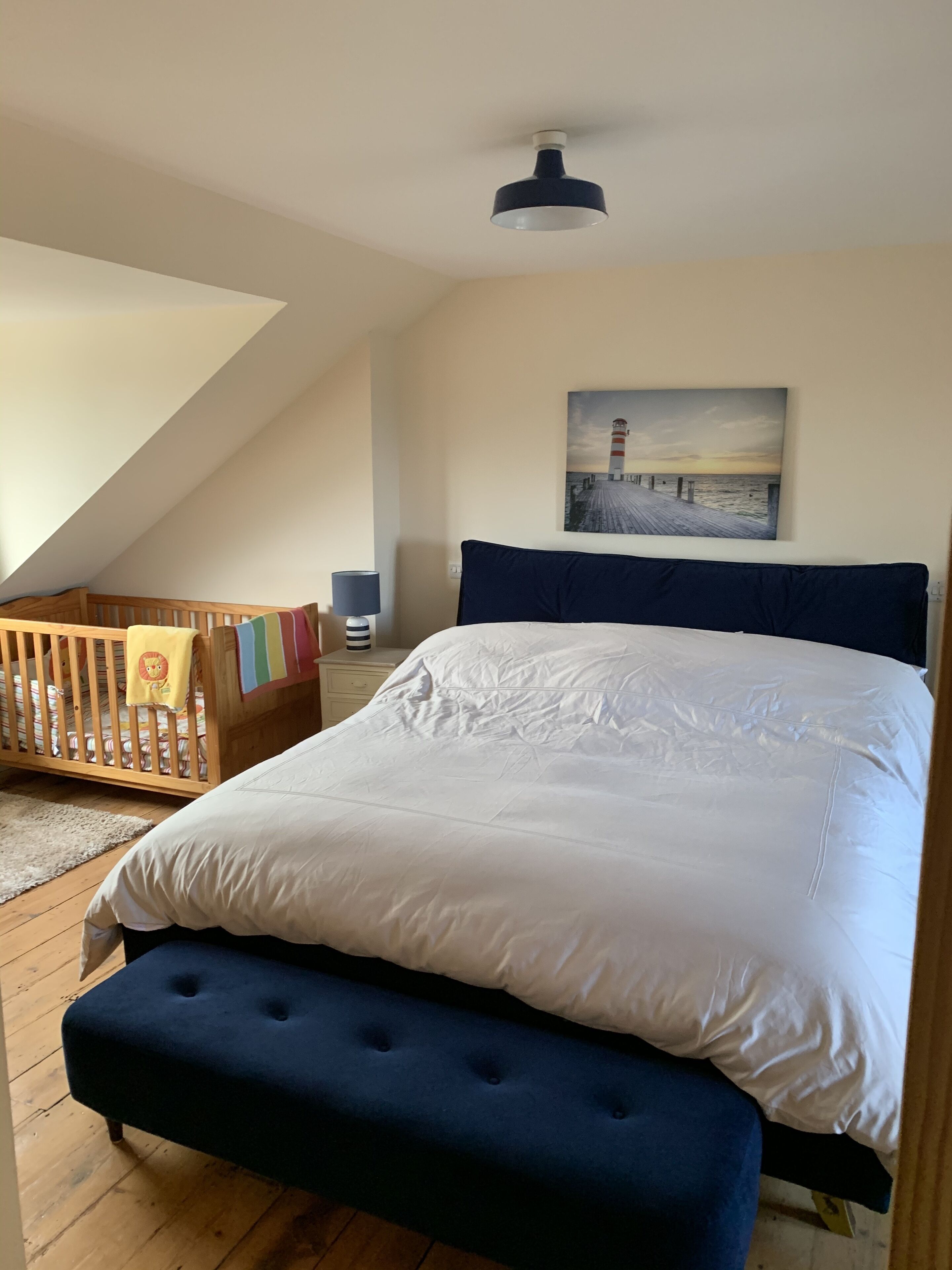 A bedroom with a large bed, a wooden crib, a painting of a lighthouse, and a sloped ceiling.