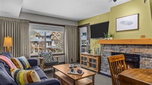 A living room with a sofa, a coffee table with wine glasses and grapes, a fireplace, a television, and a view of a mountain.