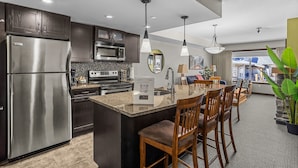 A modern kitchen with a stainless steel refrigerator, a microwave, and a built-in oven. There is a dining area with a bar stool and a large island. The kitchen has dark wooden cabinets and a stone backsplash.