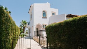 A white building with arched windows and a tiled roof, surrounded by well-maintained hedges and a paved pathway. - Cleopatra Villa (Neo Chorio)