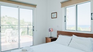 A hotel room with a bed, a bedside table with a lamp, a small drawer, and a picture on the wall. There is a balcony with chairs and a view of the outdoors. - Cleopatra Villa (Neo Chorio)