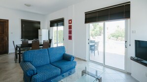 A living room with a blue sofa, a glass coffee table, a TV, a dining area with chairs and a table, and a sliding glass door leading to an outdoor patio. - Cleopatra Villa (Neo Chorio)
