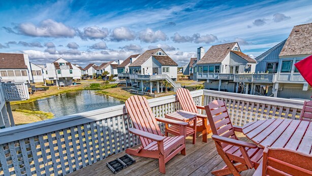 A wooden deck with Adirondack chairs and a view of houses across a body of water.