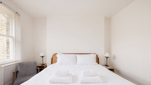 Un lit bien fait, avec du linge de lit blanc, et deux serviettes pliées au pied du lit. On y trouve également un fauteuil gris et deux tables de chevet avec des lampes.