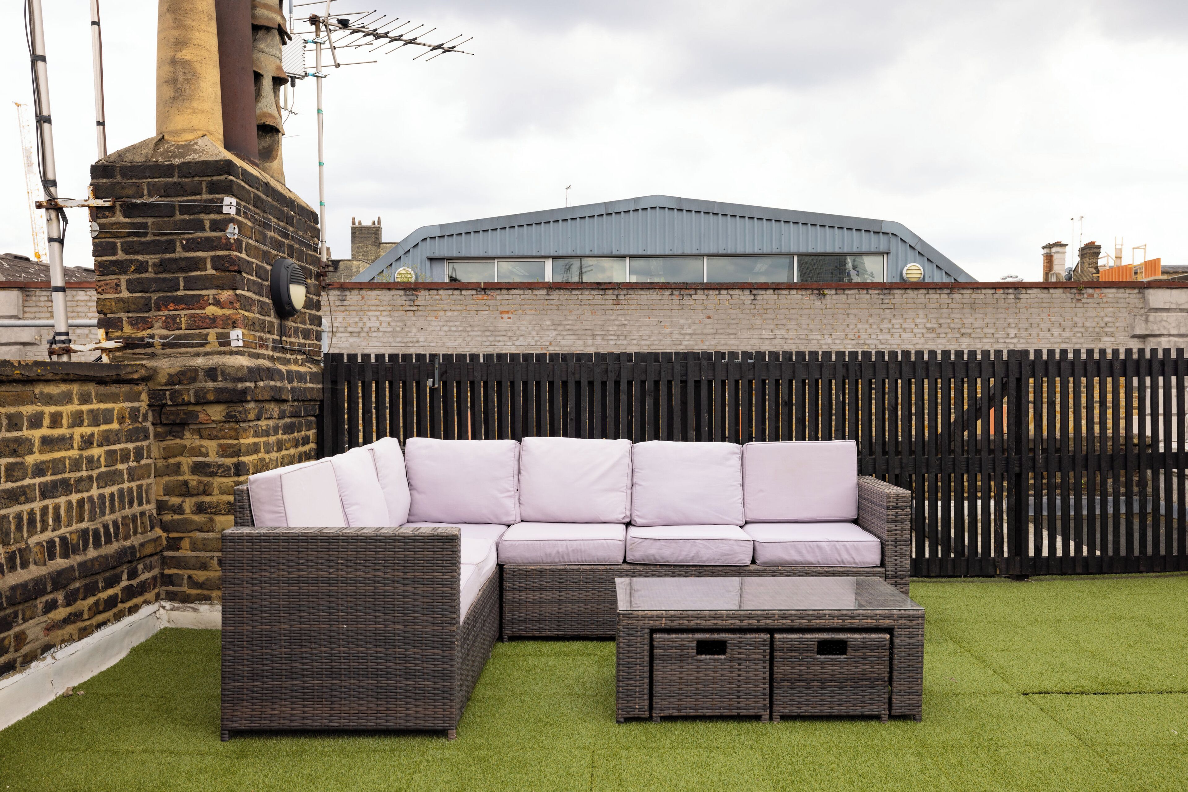 A patio area with a wicker sofa and ottoman, a brick chimney, and a metal roofed building in the background.