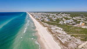 Vista aérea de un pueblo costero con una playa larga y arenosa, edificios residenciales y una carretera que sigue el curso de la costa.