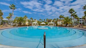 Una gran piscina cristalina con una fuente central, rodeada de palmeras y sillones de descanso, con edificios de hotel al fondo.