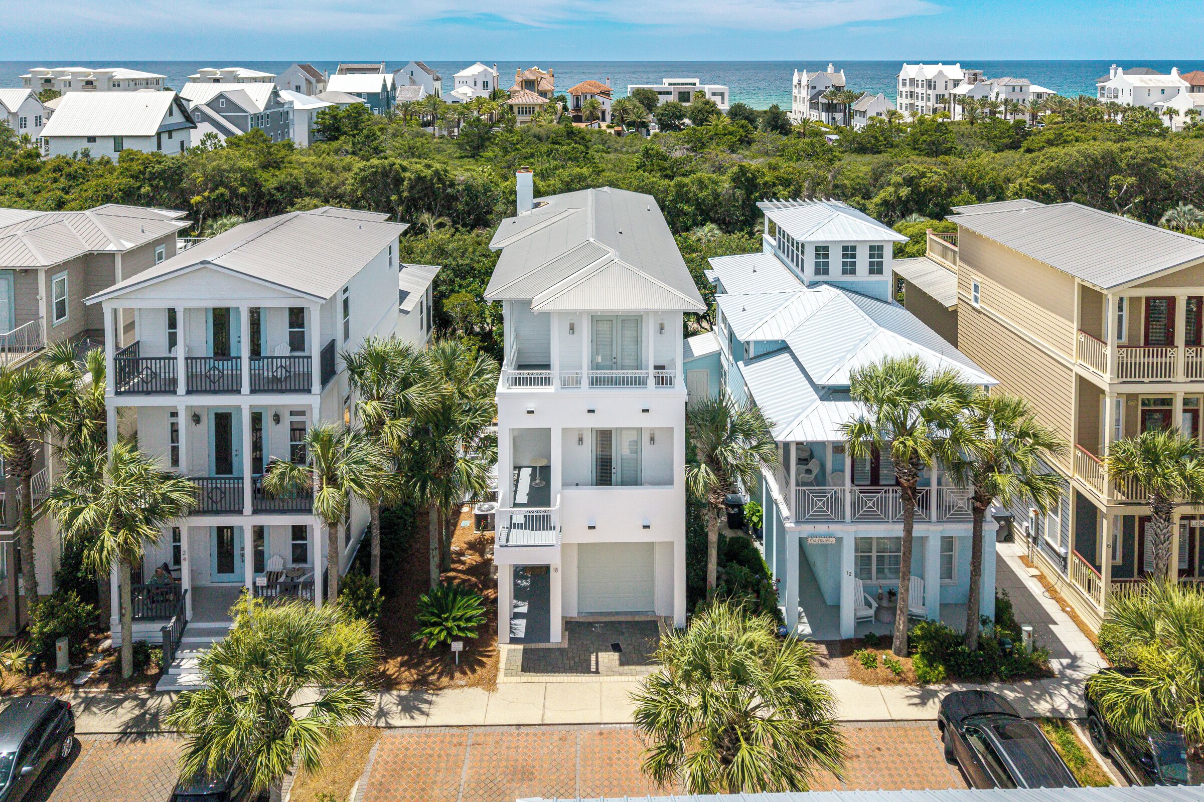 A coastal residential area with multi-story houses, some with balconies, surrounded by palm trees and a view of the ocean.