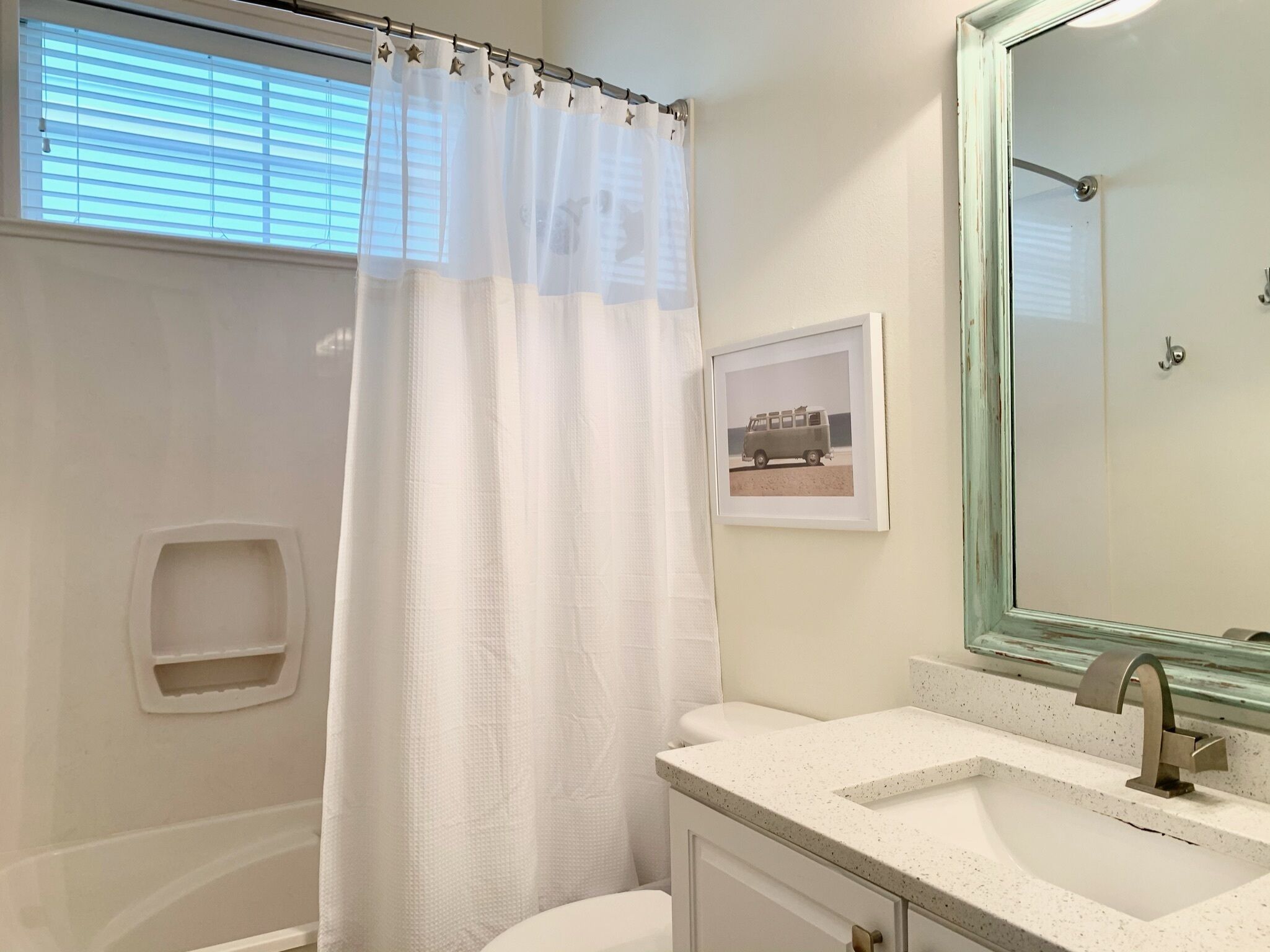 Un baño con cortina de ducha blanca, un cuadro en la pared, un lavabo con espejo y un inodoro.