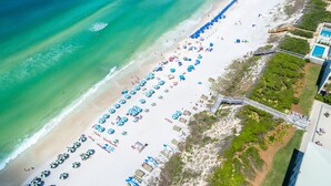 An aerial view of a beach with rows of umbrellas and beach chairs, a wooden walkway, and a pool area.