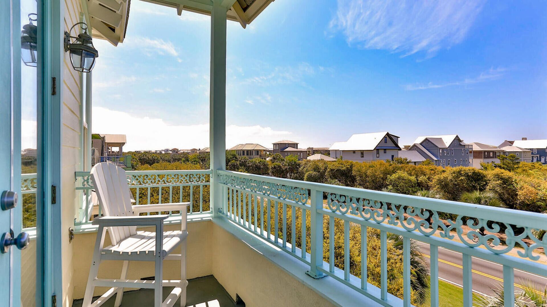 A balcony with a white chair and a small table, overlooking a residential area with houses and greenery.
