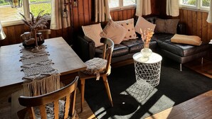 Una habitación acogedora con paredes y techo de madera, un sofá de cuero negro, una mesa de comedor de madera con sillas, y ventanas amplias que ofrecen vistas a zonas verdes y edificios.