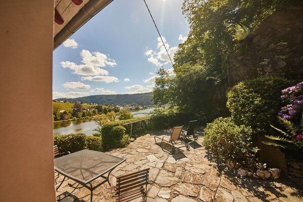 A patio with stone flooring, a table, and chairs, overlooking a river and hills.