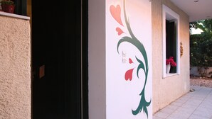 A building exterior with a textured wall, a decorative floral mural, a window with a potted plant, and a door. - Mandola Studios - Garden view Studio (Kefalonia)