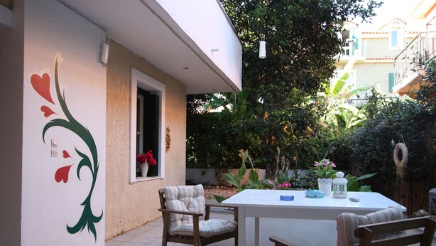 An outdoor patio with a white table and four wooden chairs, surrounded by a garden with various plants and flowers. - Mandola Studios - Garden view Studio (Kefalonia)