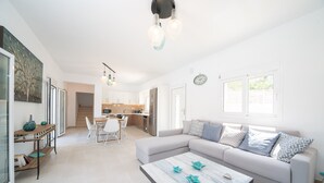 Een moderne woonkamer met een bank, salontafel en een eethoek. In de achtergrond bevindt zich een keuken met kastjes en een koelkast.