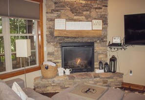 A stone fireplace with a fire, a flat-screen TV mounted on the wall, and a cozy seating area with cushions. - Charming Cabin in Deep Creek (Swanton)