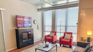A modern living room with a fireplace, a flat-screen TV mounted above it, two red armchairs, a coffee table, and a sofa. - Apartment near Kerrytown Concert House (Ann Arbor)