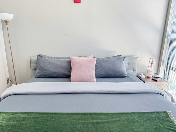 Una cama bien hecha con ropa de cama gris y una manta verde, una lámpara de noche y una mesita de noche con lámpara y control remoto.