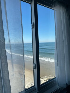 A window with sheer curtains, a view of the ocean, and a sandy beach.