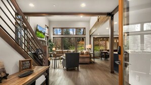 Amplia sala de estar con escalera de madera, chimenea y grandes ventanales con vistas al exterior.