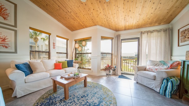 A spacious living room with a wooden ceiling, white sofas, a coffee table, and large windows with blinds. - End-Unit, 1BR/1BA, Ocean Mile J-13 'Picture Perfect', Near State Park (St. George Island)
