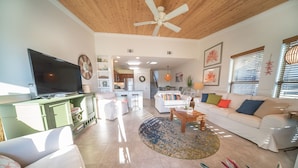A spacious living room with a television, a green cabinet, a white sofa with colorful cushions, a coffee table, and a large plant. - End-Unit, 1BR/1BA, Ocean Mile J-13 'Picture Perfect', Near State Park (St. George Island)