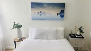 Un letto rifatto con lenzuola bianche, un dipinto di un paesaggio tranquillo sopra, un comodino con una lampada e un vaso di fiori, e una pila di asciugamani bianchi sul letto.