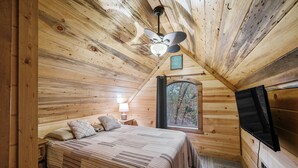 A cozy cabin bedroom with a wooden ceiling, a bed with a striped blanket, a nightstand with a lamp, and a window with a view of trees. - Dancing in the Moonlight - Walking Distance to New CASINO (Broken Bow)