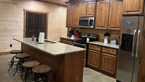 A kitchen with wooden cabinets, granite countertops, a stainless steel refrigerator, a microwave, a stove, and bar stools. - Dancing in the Moonlight - Walking Distance to New CASINO (Broken Bow)