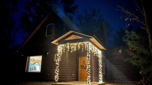 A log cabin with a porch decorated with string lights, a wooden door, and a window displaying a vehicle. - Dancing in the Moonlight - Walking Distance to New CASINO (Broken Bow)