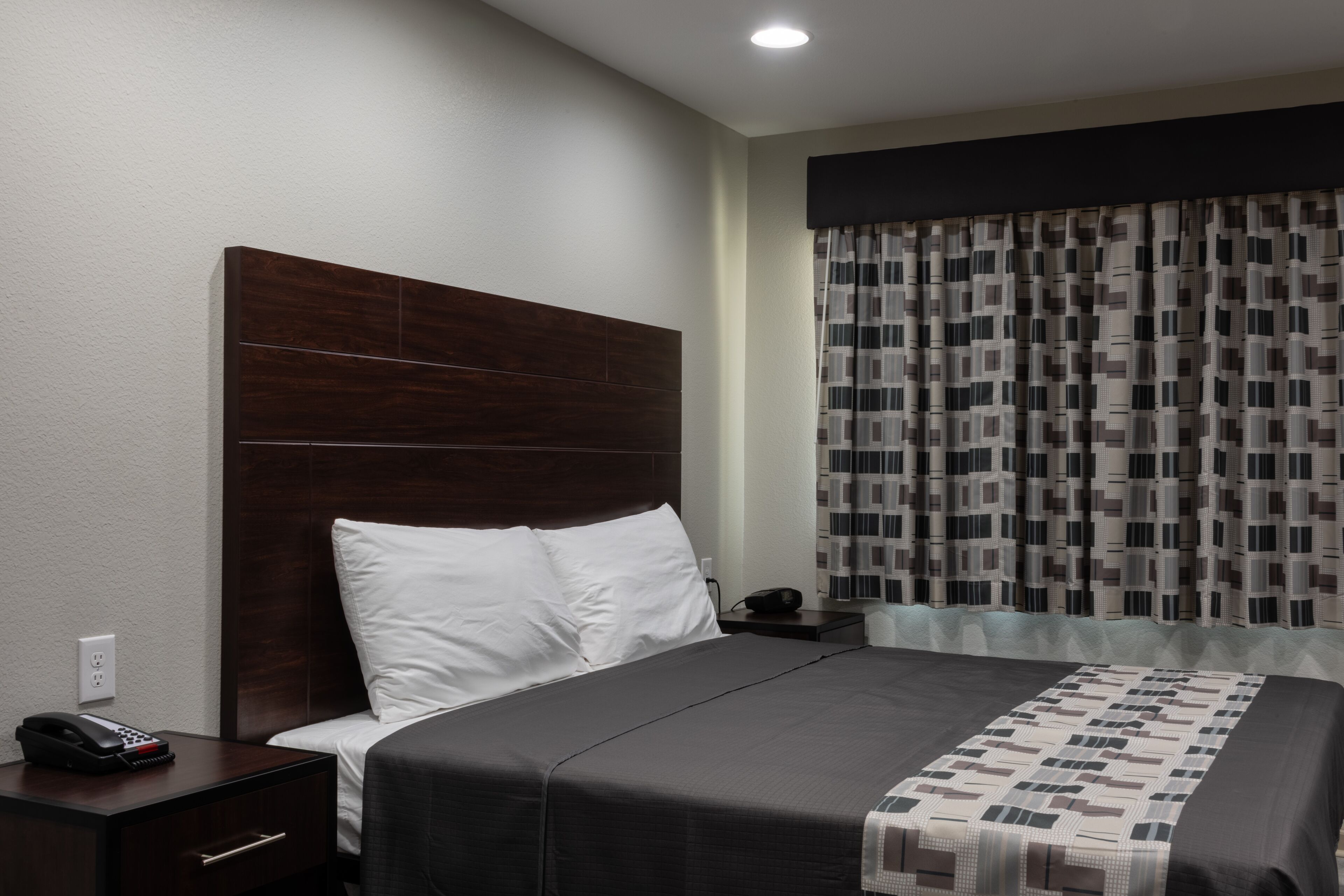 A hotel room with a large bed, a wooden headboard, a bedside table with a phone, and a patterned curtain.