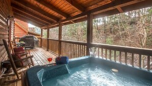 A wooden deck with a hot tub, outdoor seating, and a view of the surrounding forest.