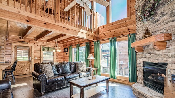 A log cabin interior with a wooden ceiling, a stone fireplace, a leather sofa, and a dining area.