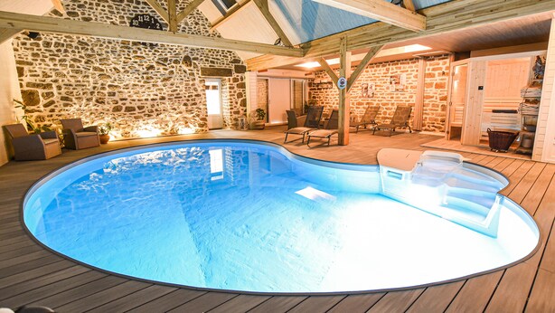 Una piscina interior con paredes de piedra y vigas de madera, rodeada de sillas de descanso y una pequeña zona de estar.