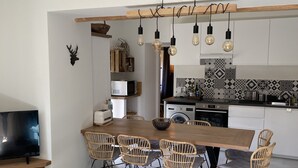 A modern kitchen with a wooden dining table, wicker chairs, and a TV on a wooden stand. - Village house, sleeps 8.  (Saint-Chaffrey)