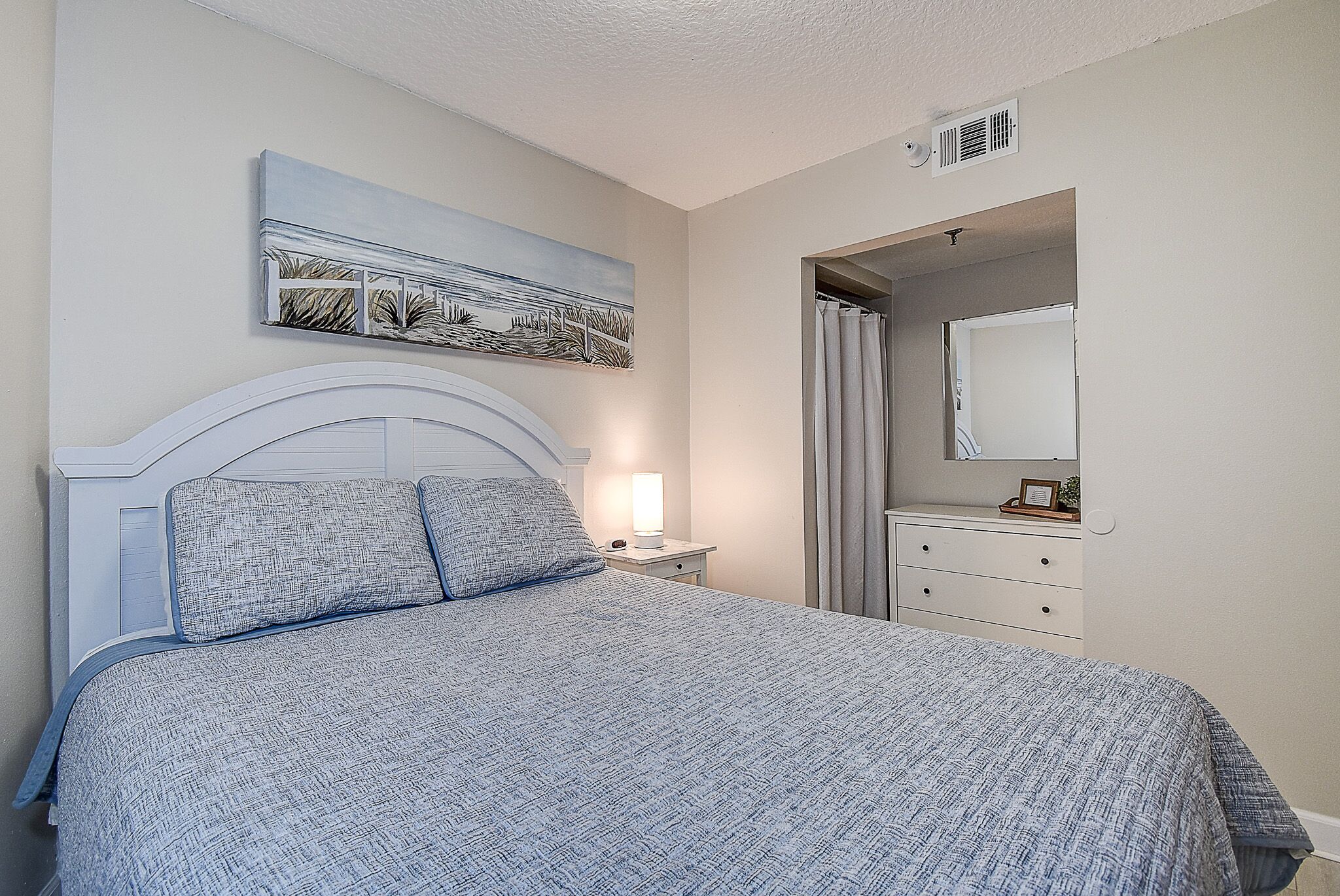 A bedroom with a bed, a nightstand, a bedside lamp, a dresser, a mirror, and a painting on the wall.
