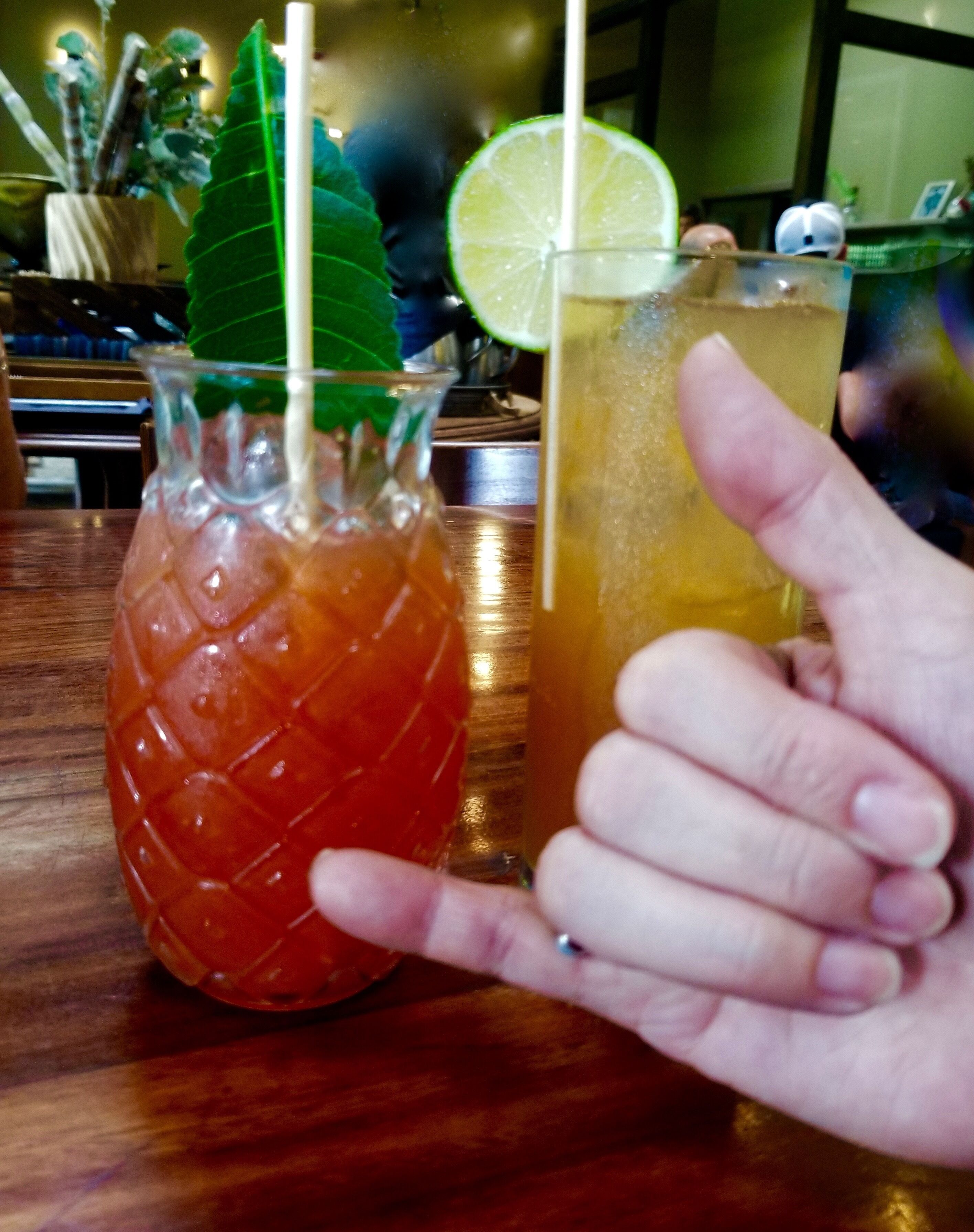 En hånd holder et glas med et limebåd, ved siden af et glas formet som en ananas med et sugerør.