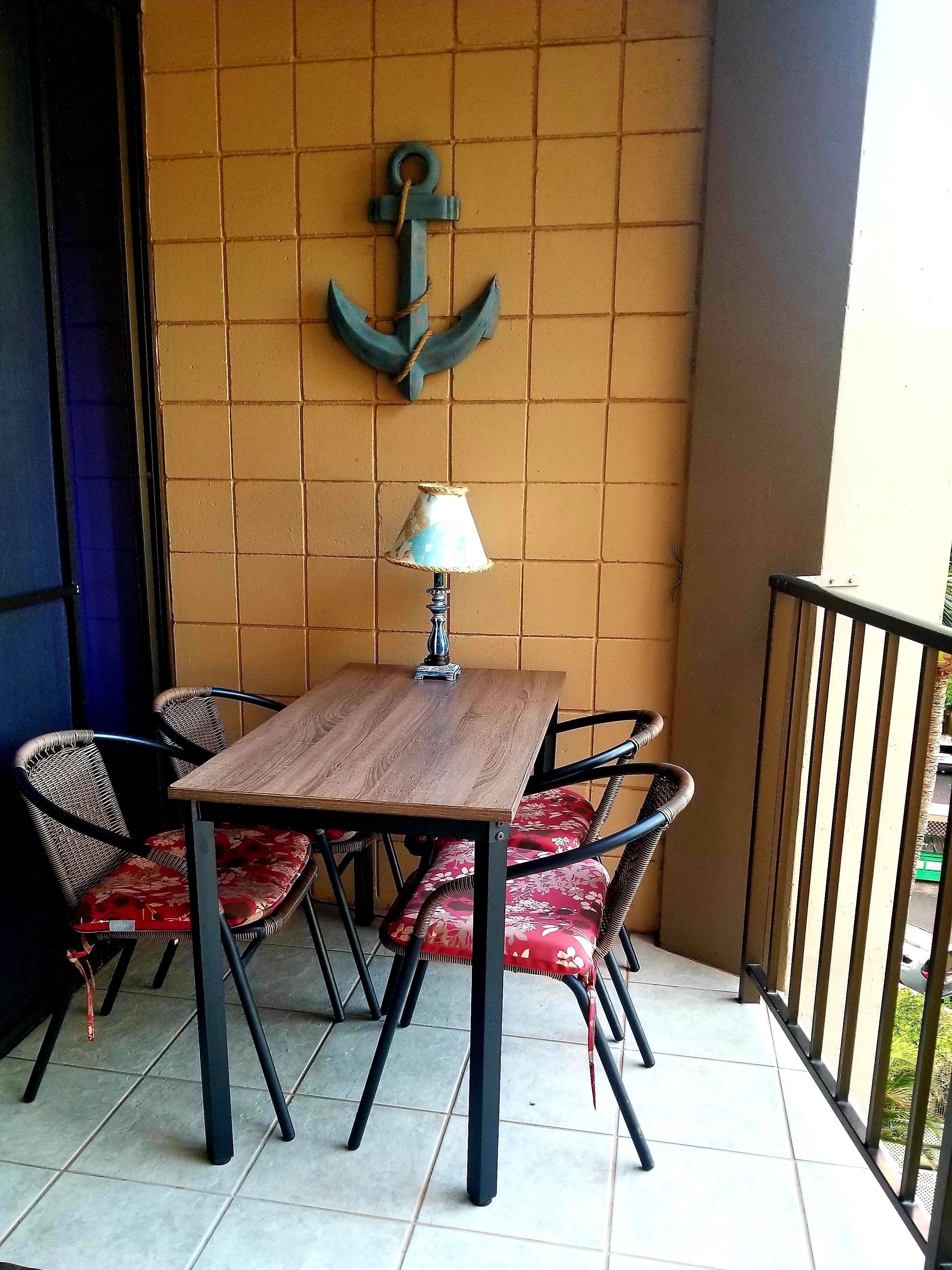 A small outdoor seating area with a wooden table and four wicker chairs with red cushions. An anchor decoration hangs on the wall behind the table.
